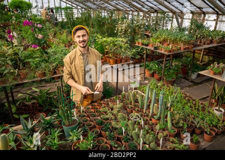Portrait of happy handsome young man in cap using small trowel while working with cactus plants in spacious greenhouse Stock Photo