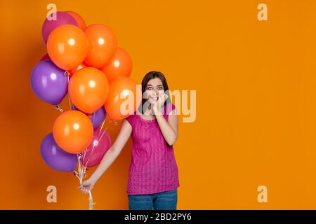 shy and embarrassed woman in pink t-shirt posing with bright colorful air balloons isolated over orange background. birthday party. space for text Stock Photo