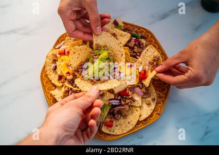 People hands dipping Yellow corn nacho chips garnished with ground beef, guacamole, melted cheese, peppers and cilantro leaves in plate on white stone Stock Photo