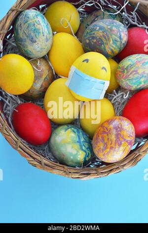 Basket with colorful eggs and a  yellow egg with protection mask on light blue background Stock Photo