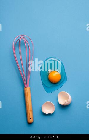 Baking and cooking concept with a kitchen utensil and broken egg. Flat lay with a cracked egg and pink whisk on a blue kitchen table. Stock Photo
