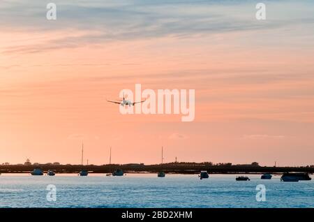 The plane flies to the airport over the river in the city of Faro in Portugal. silhouettes of airplanes and airport, boats in the river, evening city. Stock Photo