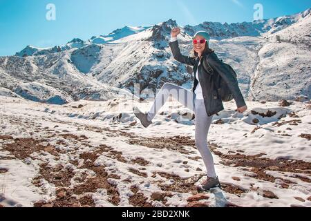 A young girl in glasses dances on the snow. Mountain peaks in the winter season. Stock Photo