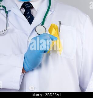 doctor in a white uniform and sterile latex gloves holds a yellow ribbon in his hand, symbol of the fight against the cat's tumor, problem of suicides Stock Photo