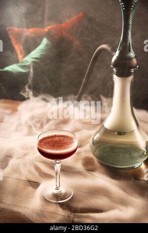 A glass of raspberry cocktail next to a hookah on a wooden table in a dark bar. Alcohol and smoking concept. Stock Photo