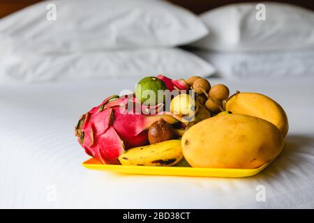 Compliment surprise plate of fruits in bedroom. Exotic fruits on tray at home, hotel resort white bed sheet with mango, pitaya dragon fruit, banana, l Stock Photo