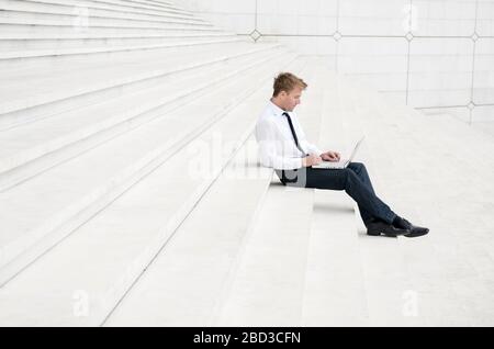Young man in a smart casual white shirt and jeans sitting outdoors on minimalist marble steps using his laptop computer Stock Photo