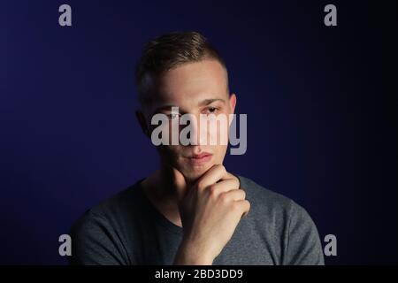 Young caucasian blonde man in grey t shirt on dark purple background touching his chin and thinking about something Stock Photo