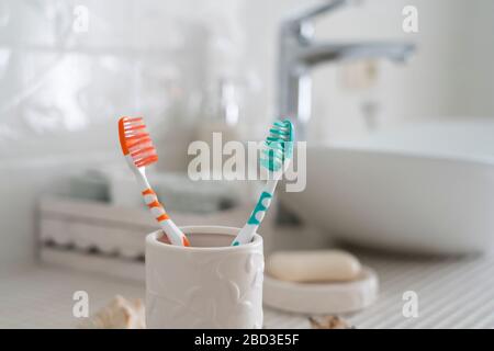 Two colorful toothbrushes stand in a ceramic glass against the background of the washbasin and other accessories in the bathroom. Stock Photo