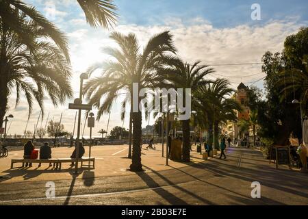 Backlit view of the waterfront of the coastal town of Riviera of Flowers with people walking and sitting on benches, Arma di Taggia, Liguria, Italy Stock Photo
