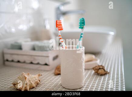 Two colorful toothbrushes stand in a ceramic glass against the background of the washbasin and other accessories in the bathroom. Stock Photo