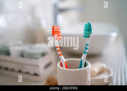 Two colorful toothbrushes stand in a ceramic glass against the background of the washbasin and other accessories in the bathroom. Stock Photo