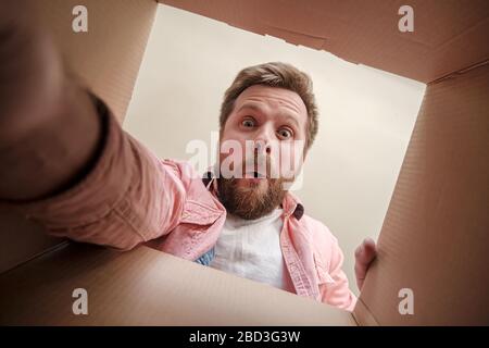 Satisfied, surprised man holds out his hand, trying to get a gift or parcel from an unpacked box. Unboxing inside view. Stock Photo