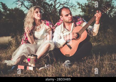 Romantic couple hugging while having picnic in sunset light. Man with guitar and attractive young woman in white dress are sitting together on grass Stock Photo