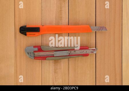 Two stationery knives in orange and red on a wooden background. Office supplies top view. Old stationery knives with narrow blades. Stock Photo
