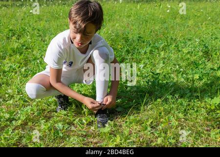 Close up view portrait of little boy in white soccer uniform trying to tie lace on his sportshoe on green grass field. Young sportsman, future champio Stock Photo