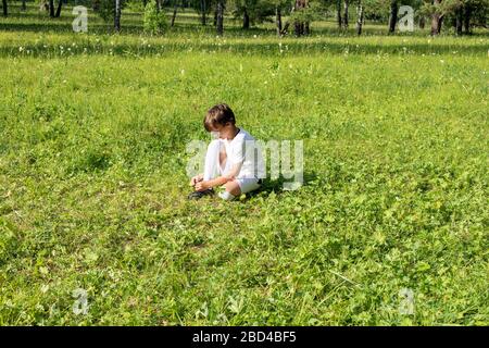 Close up view portrait of little boy in white soccer uniform trying to tie lace on his sportshoe on green grass field. Young sportsman, future champio Stock Photo