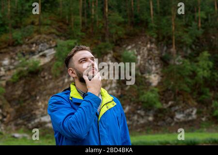 Close up portrait of handsome young man looking up thoughtfully touching beard with hand on blurred natural background. Nowhere to hurry. Solving prob Stock Photo