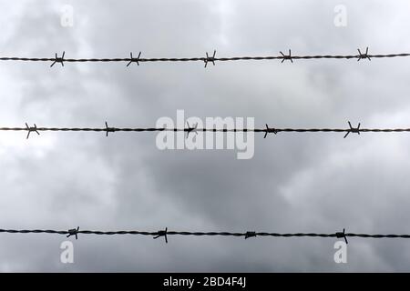 Barbed wire background. Three horizontal lines of barbed wire against a dramatic dark cloudy sky in monochrome colors. No access or trespassing. Stock Photo