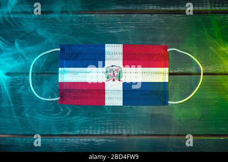 Medical mask for protection against airborne diseases, painted in the national flag of Dominicana  on a black background in blue-green smoke. Medical Stock Photo