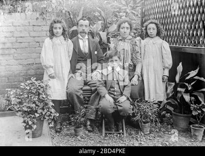 A vintage early Edwardian black and white p;photograph taken in England, showing a family, mother, father, two daughters, and a son. They are posing for the photographer in their back yard, or garden, and are dressed in their best fashionable clothes. Stock Photo