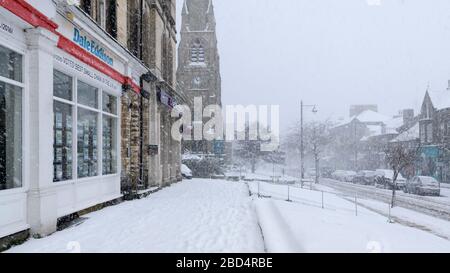 Snowy winter townscape (snowing, snow-covered high street, church, shops & road in scenic town centre) - The Grove, Ilkley, Yorkshire, England, UK. Stock Photo