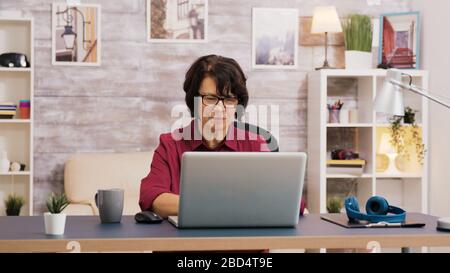 Elderly age woman taking a sip of coffee while browsing on laptop. Old man relaxing on sofa in the background. Stock Photo