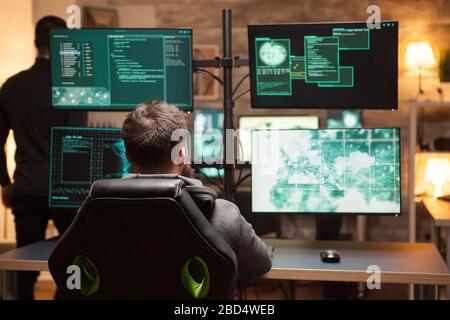 Rear view of hacker in front of computer with multiple screens in dark room. Stock Photo