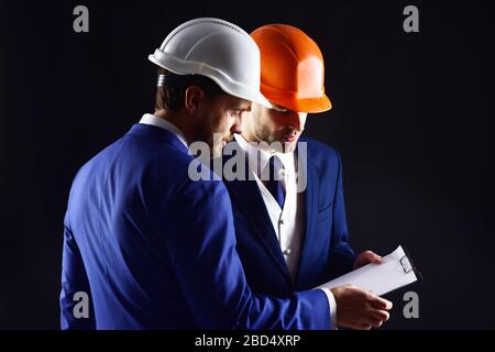 Engineering, business concept. Business partners in helmets with serious faces. Business partners look at plan. Architects discuss project. Entrepreneur consults with architect about building plans. Stock Photo