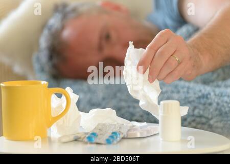 Man suffering from flu or cold lies sadly covered with a blanket on his bed. Drugs, medicines, cup and tissues in foreground Stock Photo