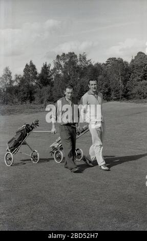 1960s, historical, Outside on an inland tree-lined golf course, two male golfers walking up a fairway together pulling their golf bags on trolleys or karts of the era, England, UK. Stock Photo