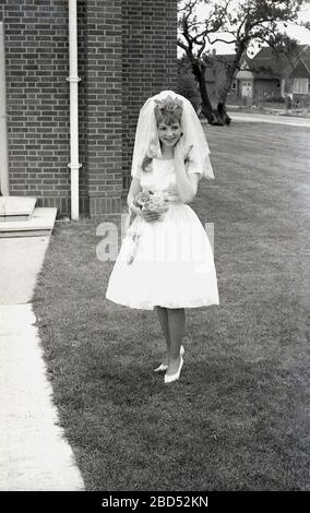 1960s, historial, teenage bride in her wedding dress standing outside the modern church building for a photograph, England, UK.  In this era as in previous ones, people married at far younger age than later generations. Stock Photo