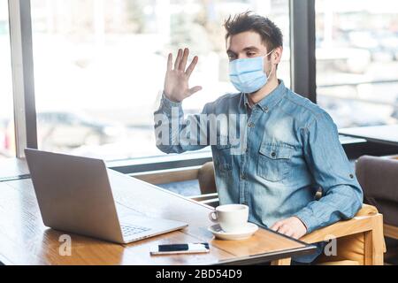 Portrait of satisfied happy young businessman with surgical medical mask in blue shirt sitting and greeting a worker through a webcam on online meetin Stock Photo