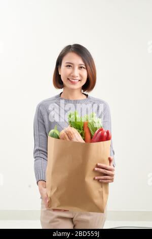 Beautiful smiling Asian woman holding paper shopping bag full of vegetables and groceries, studio shot isolated on white background Stock Photo