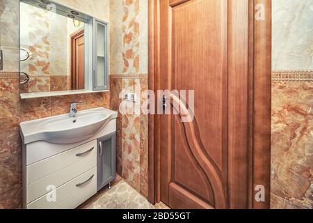 Small beige tile bathroom with sink and mirror and brown wooden door Stock Photo