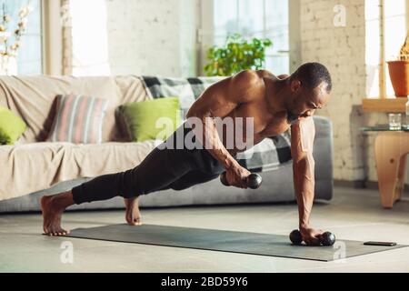 African-american man teaching at home online courses of fitness, aerobic, sporty lifestyle while being quarantine. Getting active while isolated, wellness, movement concept. Training with weights. Stock Photo