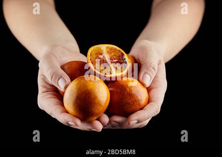 Red or blood oranges in hands on black background. Person holging handful of whole and halved oranges. Heap of unpeeled citrus fruits, selective focus Stock Photo