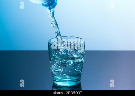 In a glass standing on a black table pour clean water from a bottle. Blue glass with flowing water standing on a black table on a blue background Stock Photo
