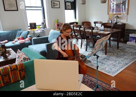 New York, USA. 2020. Schoolgirl learning to play a Cello using a laptop  to watch and listen her music teacher from her apartment during the Coronavir Stock Photo