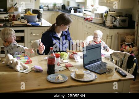 Mother feeding her children dinner at kitchen table whilst working from home during period of self-isolation - 2020 COVID-19 coronavirus pandemic Stock Photo