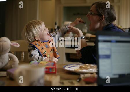 Mother feeding her children dinner at kitchen table whilst working from home during period of self-isolation - 2020 COVID-19 coronavirus pandemic Stock Photo