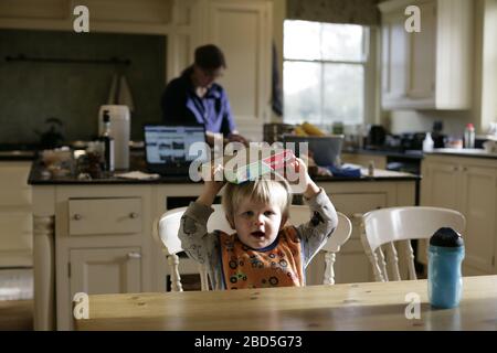 Child toddler playing with jigsaw puzzle whilst mother cooking dinner in kitchen during period of self-isolation - 2020 COVID-19 coronavirus pandemic Stock Photo
