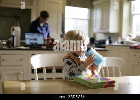 Child toddler drinking from dinks bottle whilst mother cooking dinner in kitchen during period of self-isolation - 2020 COVID-19 coronavirus pandemic Stock Photo