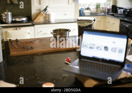 Mother prepares family dinner at home whilst working on laptop in kitchen during self-isolation during the 2020 COVID-19 coronavirus pandemic Stock Photo