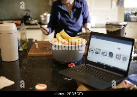 Mother prepares family dinner at home whilst working on laptop in kitchen during self-isolation during the 2020 COVID-19 coronavirus pandemic Stock Photo