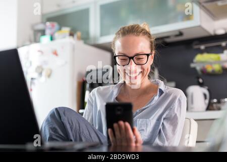 Stay at home and social distancing. Woman in her casual home clothing working remotly from kitchen dining table. Video chatting using social media
