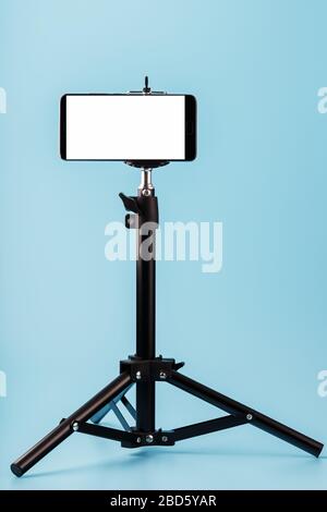 Mobile phone mounted on a tripod with a white display free for images and text, blue isolated background. Stock Photo