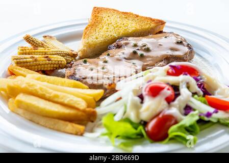 Closeup side view steak pork chop with creamy pepper sauce, Served with french fries, toast, grill baby corn, roast carrot, and vegetables salad on a Stock Photo