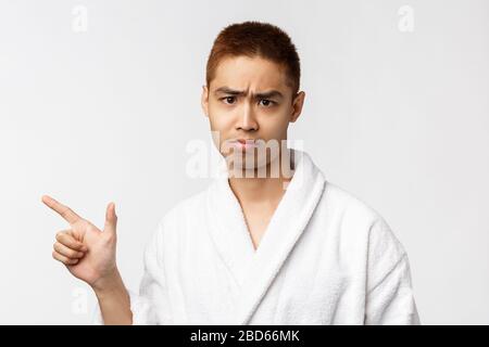 Beauty, spa and leisure concept. Portrait of skeptical, upset and moody asian man in bathrobe, sighing and frowning, sulking disappointed, pointing Stock Photo