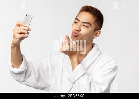 Beauty, spa and technology concept. Portrait of cheeky young handsome asian man in bathrobe, show kissing face at mobile camera, taking selfie Stock Photo